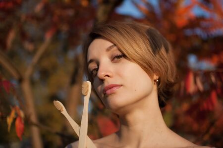 Young girl with short blond hair is holding a bamboo toothbrushes outdoors during sunny weather in...