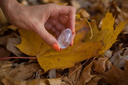 Female hand with orange manicure holding transparent violet amethyst yoni egg for vumfit, imbuilding or meditation on yellow fallen leaves background during autumn day outdoorsの写真素材