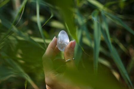 Female hand with french manicure holding transparent violet amethyst yoni egg for vumfit, imbuilding or meditation. Crystal quartz egg in hands on green background outdoorsの写真素材