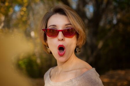 Emotional attractive young woman with blonde short hair, biege dress and red fashoinable sunglasses grimacing in a beautiful park outdoors in autumnの写真素材