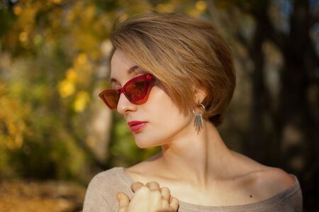 Young and beautiful girl with short hair and red fashionable sunglasses is posing against the trees with yellow leaves background spending time in the autumn parkの写真素材