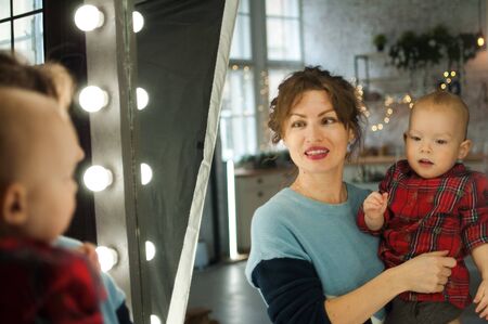 Young beautiful mother in blue sweater holding her son in red shirt and looking at the mirror at home in the room with Christmas decoration, Xmas tree and lightsの写真素材