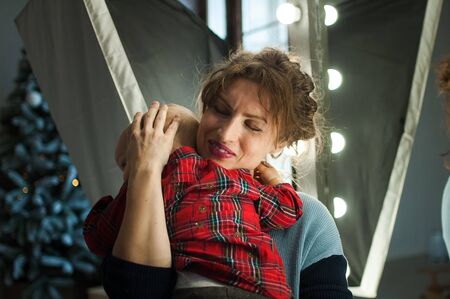 Attractive curly young mother in blue sweater hugging her toddler baby in red shirt in the room with Christmas decoration, Xmas tree and lightsの写真素材