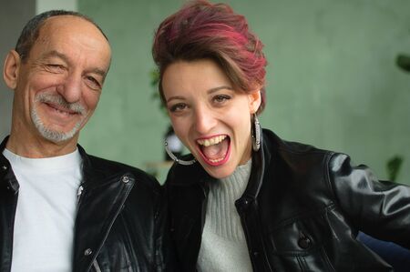 Indoor family portrait of adult daughter and senior father in loft room with houseplants. Crazy man and girl with pink hair are wearing black leather jackets in punk style and smilingの写真素材