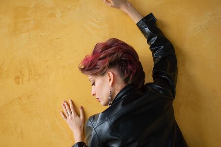 Back view of glamorous lady in punk rock style party clothes with dark pink hair in casual dress, black leather jacket, and long earrings on yellow backgroundの写真素材