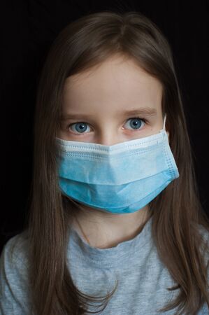 Little blond girl with blue eyes and long hair is posing on black backgound in surgical disposable mask for protection of Coronavirus Covid-19 during self-isolation while staying at homeの写真素材