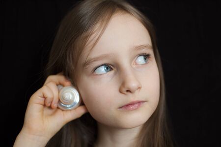 Little girl is dreaming about the summer trip to the ocean and listening to a white shell to hear a sea sound on black background in studioの写真素材