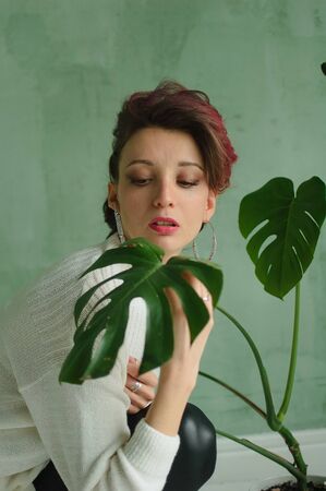 Studio portrait of young fashion model wearing white sweater and black leather leggings possing at studio with green background and exotic plantsの写真素材