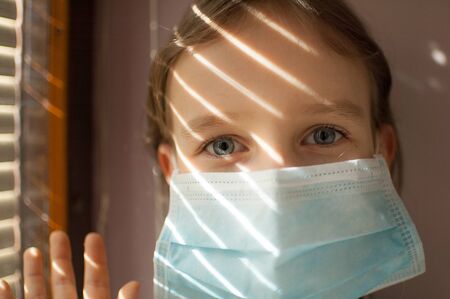 Coronavirus Covid-19 outbreak. Little blonde girl with blue eyes wearing disposable mask for protection of virus sitting near the window with blinds. Personal protective equipmentの写真素材