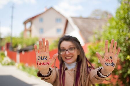 Smiling girl with long dreadlocks and eyeglasses in nude color trench is showing hands with written slogan Our future in our hands on some building background. Positive psychology conceptの写真素材