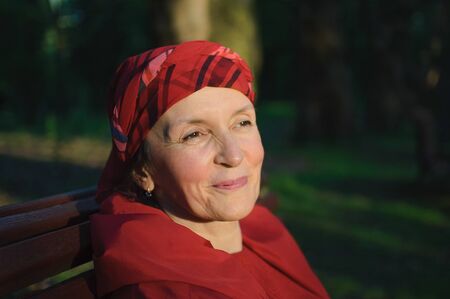 Female portrait of mature woman dressed in red clothes and sitting on the bench and enjoying a good weather and sunset in the park during a spring or autumn.の写真素材