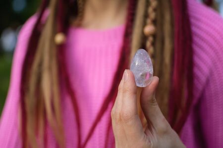 Adult girl in knitted sweater with long dreadlocks is holding transparent violet amethyst yoni egg for vumfit, imbuilding or meditation outdoors on her body background outdoorsの写真素材