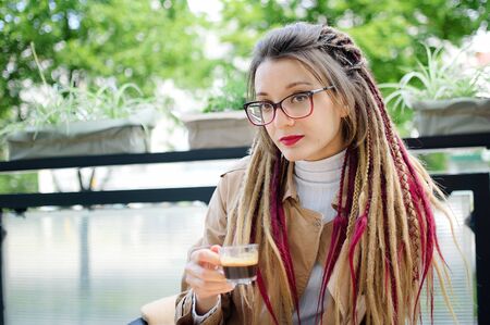 Positive smart student girl with long dreadlocks is resting in a street coffee shop with transparent cup of espresso.の写真素材