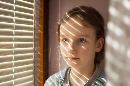 Portrait of a little girl with long blond hair and blue eyes is sitting near the window with blinds during sunny day at homeの写真素材