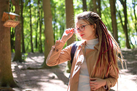 Young woman wearing glasses, casual nude trench, and dreadlocks is standing at the urban park on...