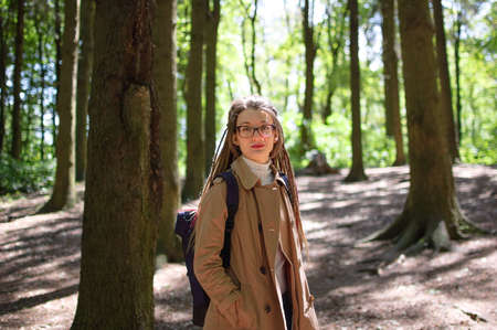 Young woman wearing glasses, casual nude trench, and dreadlocks is standing at the urban park on...