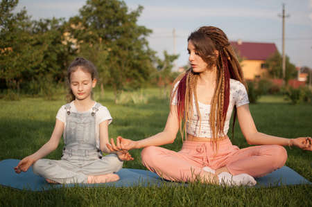 Young mother with dreadlocks and little daughter are doing yoga exercises in lotus position on grass in the park at the day time. Concept of friendly family and of summer vacation.の写真素材