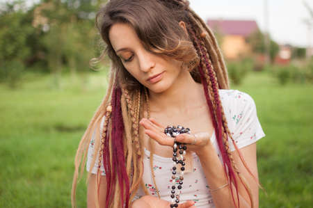 Young beautiful girl with long dreadlocks holds in hands the necklace made by natural stones for meditation outdoors during morning on green grass of her backyardの写真素材