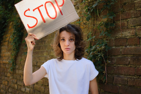 Young protesting woman in white shirt and jeans holds protest sign broadsheet placard with slogan Stop for public demonstration on wall background.の写真素材