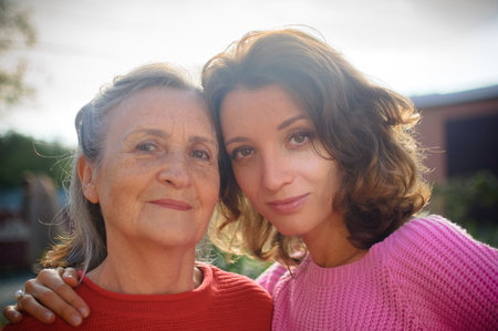 Senior mother with gray hair with her adult daughter looking at the camera in the garden and hugging...