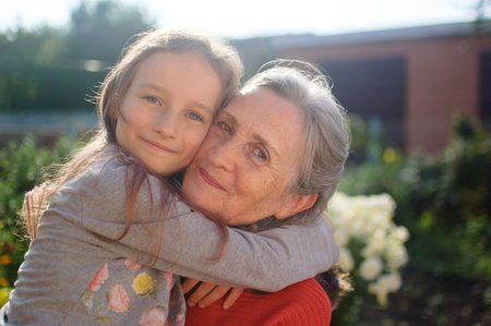 Senior grandmother with gray hair wearing red sweater with her little granddaughter are hugging in the garden and during sunny day outdoors, mothers dayの写真素材