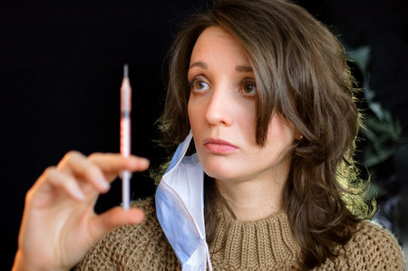 Vaccine concept. Female portrait of caucasian woman with face mask holding a syringe and looking at it on black background in studio.の写真素材
