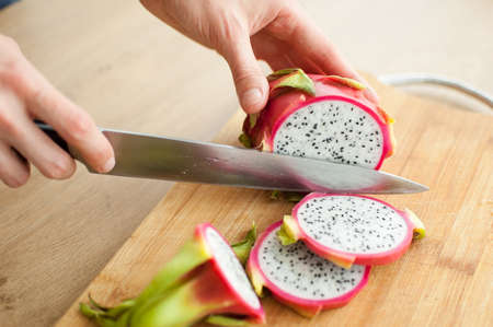 Female hands is cutting a dragon fruit or pitaya with pink skin and white pulp with black seeds on wooden cut board on the table. Exotic fruits, healthy eating conceptの写真素材