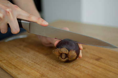 Female hands is cutting with knife a fresh ripe mangosteen on a cut wooden board. Exotic fruits,...