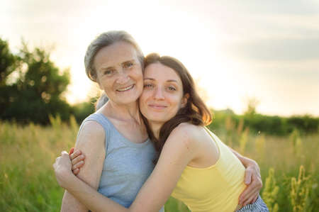 Senior mother with gray hair with her adult daughter looking at the camera in the garden and hugging...