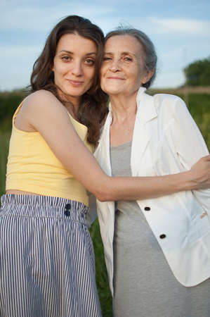 Senior mother with gray hair with her adult daughter looking at the camera in the garden and hugging...