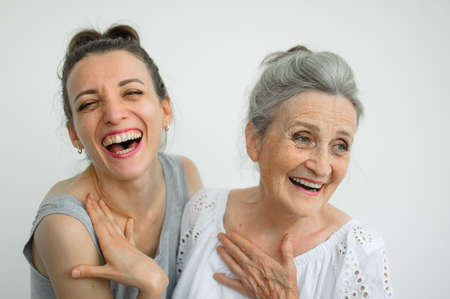 Happy senior mother is hugging her adult daughter, the women are laughing together, sincere family of different age generations having fun on white backgroundの写真素材