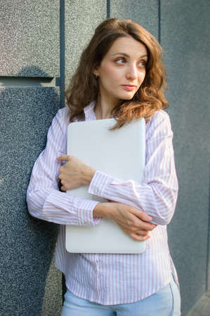 Female portrait of young woman with silver laptop, businesswoman is posing with digital tablet outside on dark wall background while waiting for a meeting during a break from workの写真素材