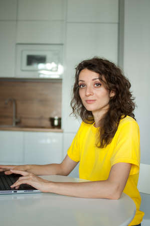 Young woman with curly hair and yellow shirt is working from home using her laptop at the kitchen...