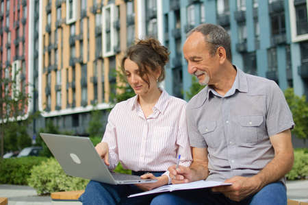 Attractive young woman and senior man are using laptop sitting on the bench in modern residential complex. Casual meeting outside office. A daughter is teaching technology for her fatherの写真素材