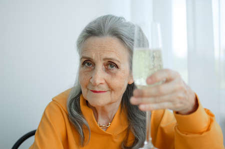 Beautiful old grandmother with grey hair and face with wrinkles sitting at the table at home on window background with glass of champagne, mothers day, happy retirementの写真素材
