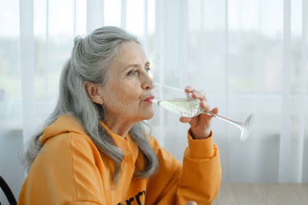 Beautiful old grandmother with grey hair and face with wrinkles sitting at the table at home on window background with glass of champagne, mothers day, happy retirementの写真素材