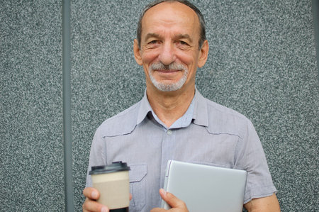 Male portrait of older coworker with silver laptop and cup of coffee waiting for a meeting near dark...