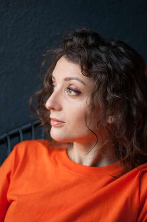 Female portrait of a beautiful curly girl in bright orange t-shirt at home in her apartment on dark background, happy people conceptの写真素材