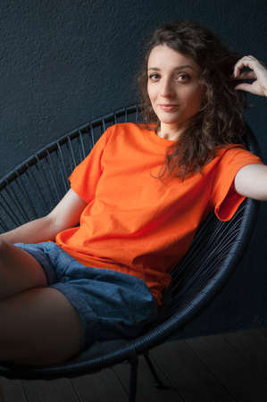 Female portrait of a beautiful curly girl in bright orange t-shirt sitting at home in her apartment on dark background, happy people conceptの写真素材