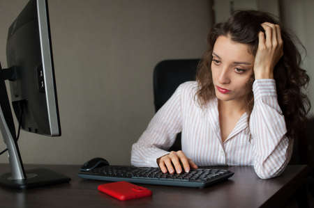 Tired young woman with curly hair and white shirt is working at the office using her laptop, routine work, freelance, burnout syndromeの写真素材