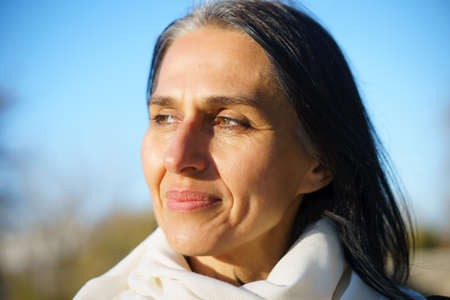 Profile side view portrait of attractive cheerful dreamy grey-haired middle-aged woman relaxing outdoors during sunny dayの写真素材