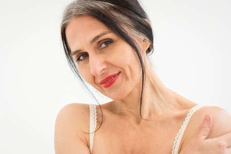 Middle aged woman starting getting grey-haired is posing in studio with naked shoulders on white background, face skin care beauty, middle age skincare cosmetics, cosmetology conceptの写真素材