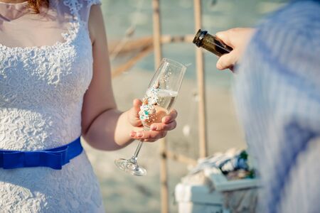 Glass with champagne is  in the bride's hands. The groom is pouring champagne into the bride's glass.  Sea style wedding.の写真素材