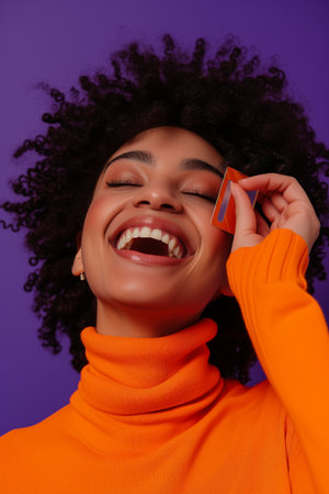 Cheerful young african american woman holding credit card and laughing while standing against purple backgroundの素材