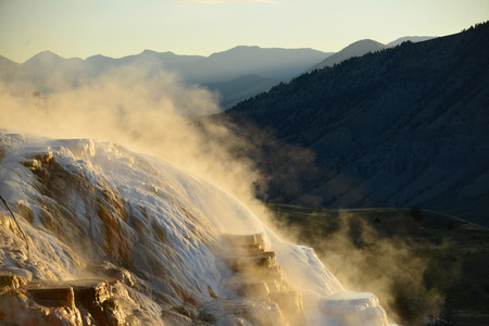 Geothermal flow of hot, carbonate rich water, forms cascading, dark orange travertine terraces, with steam rising and mountains in the background, at Mammoth Hot Springs in Yellowstone, Wyomingの写真素材