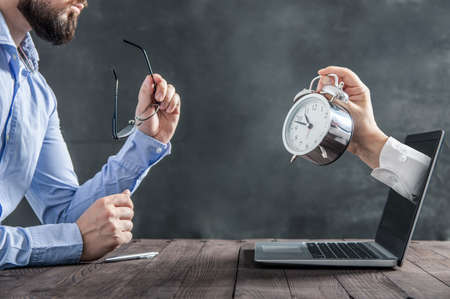 Businessman is sitting at the desk and is looking at the hand with clock coming out of the laptop....
