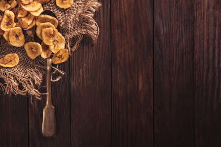 Dried bananas stacked on a fabric and spoon over old brown table. Composition in the old style with a view from the top and copy space for your textの写真素材