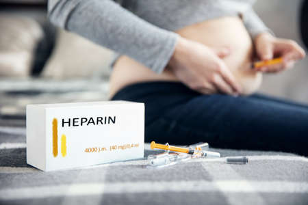 Syringes with medicine and a box with "heparin" inscription lies on the blanket. Pregnant woman makes an injection in the background. Inscriptions on the box made in graphic program.の写真素材