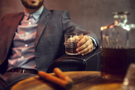 Elegant man with the glass of whisky and two cigars and a carafe of whisky on wooden table in the foregroundの写真素材