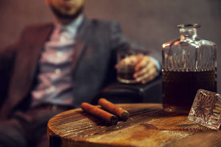 Two cigars and a carafe of whisky on wooden table and an elegant man with the glass of whisky in the backgroundの写真素材